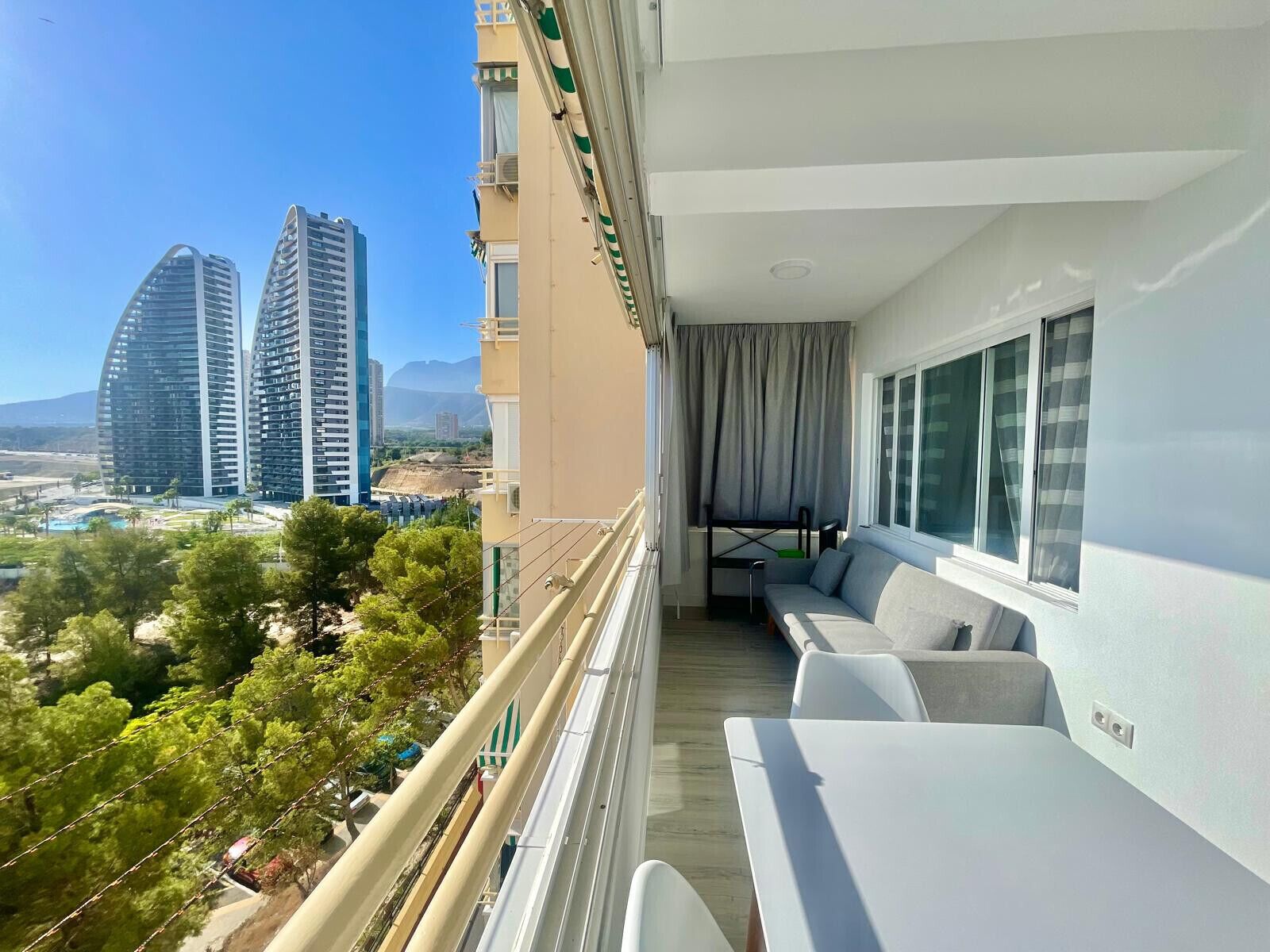 A balcony with a sofa and a dining table, overlooking a cityscape with modern buildings and a pool.