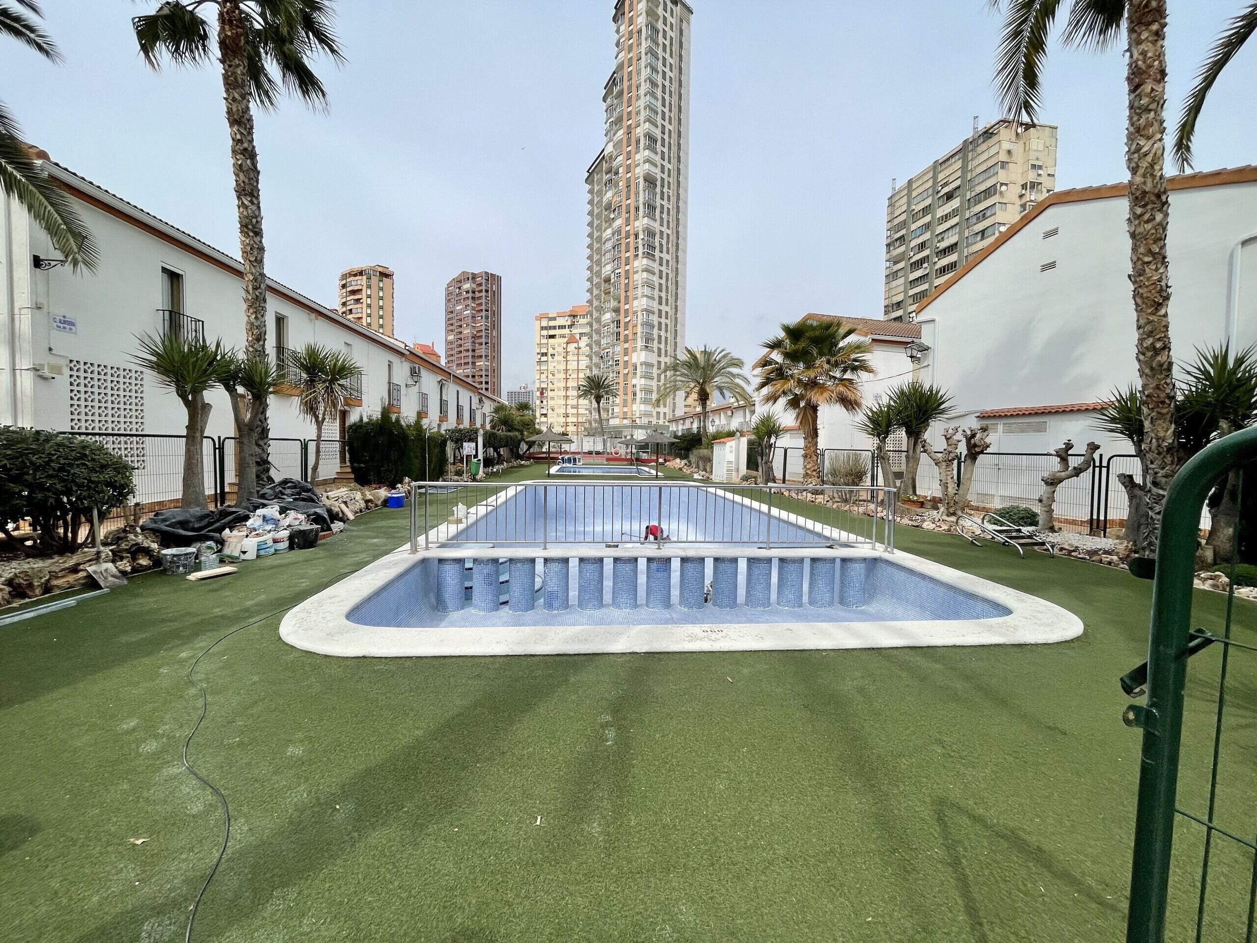 A swimming pool with a surrounding green lawn, palm trees, and tall residential buildings in the background.