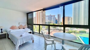 A hotel room with a bed, a small table, and chairs, a large window with a city view, and a balcony with a pool.