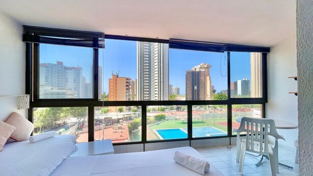 A hotel room with a large bed, a chair, and a view of a cityscape through large windows.