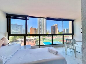Habitación de hotel con una cama grande, una silla y vistas de un paisaje urbano a través de amplios ventanales.