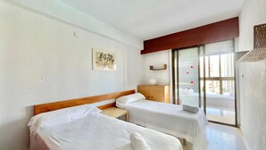 A hotel room with two single beds, a wooden headboard, a small bedside table, a wall-mounted artwork, and a sliding glass door leading to a balcony.