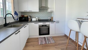 A modern kitchen with white cabinets, a stainless steel sink, a built-in oven, and a small dining area with a white chair.