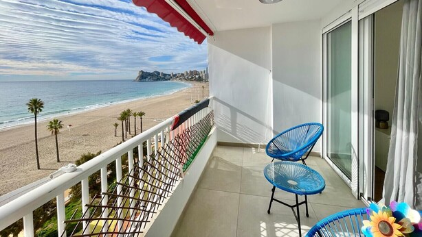 A balcony with a view of the beach, a blue chair, and a white railing.
