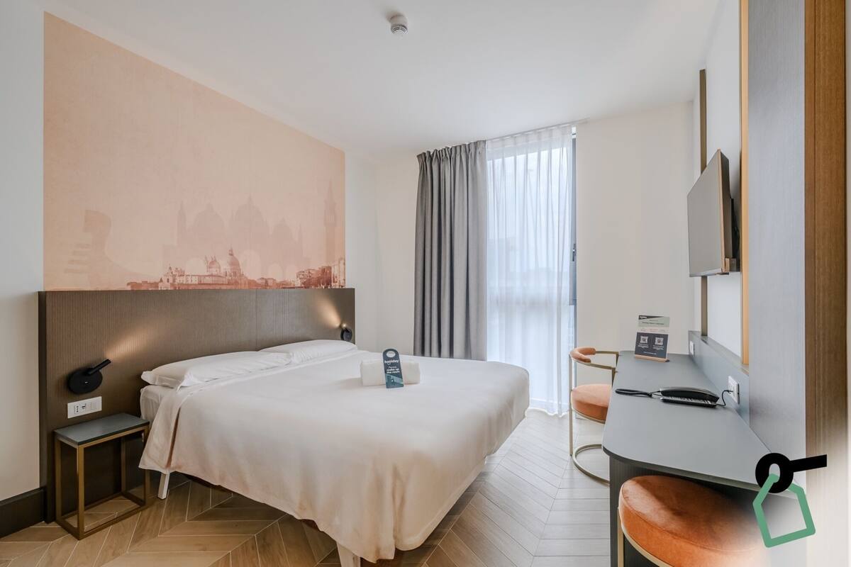 A hotel room with a bed, bedside table, lamp, and a desk with a chair.