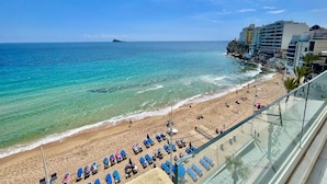 A beach with clear turquoise waters, a sandy shore, and a rocky cliff. There are blue lounge chairs and umbrellas along the beach. - EMALCO SEAFRONT MODERN APARTMENT A061 (Benidorm)