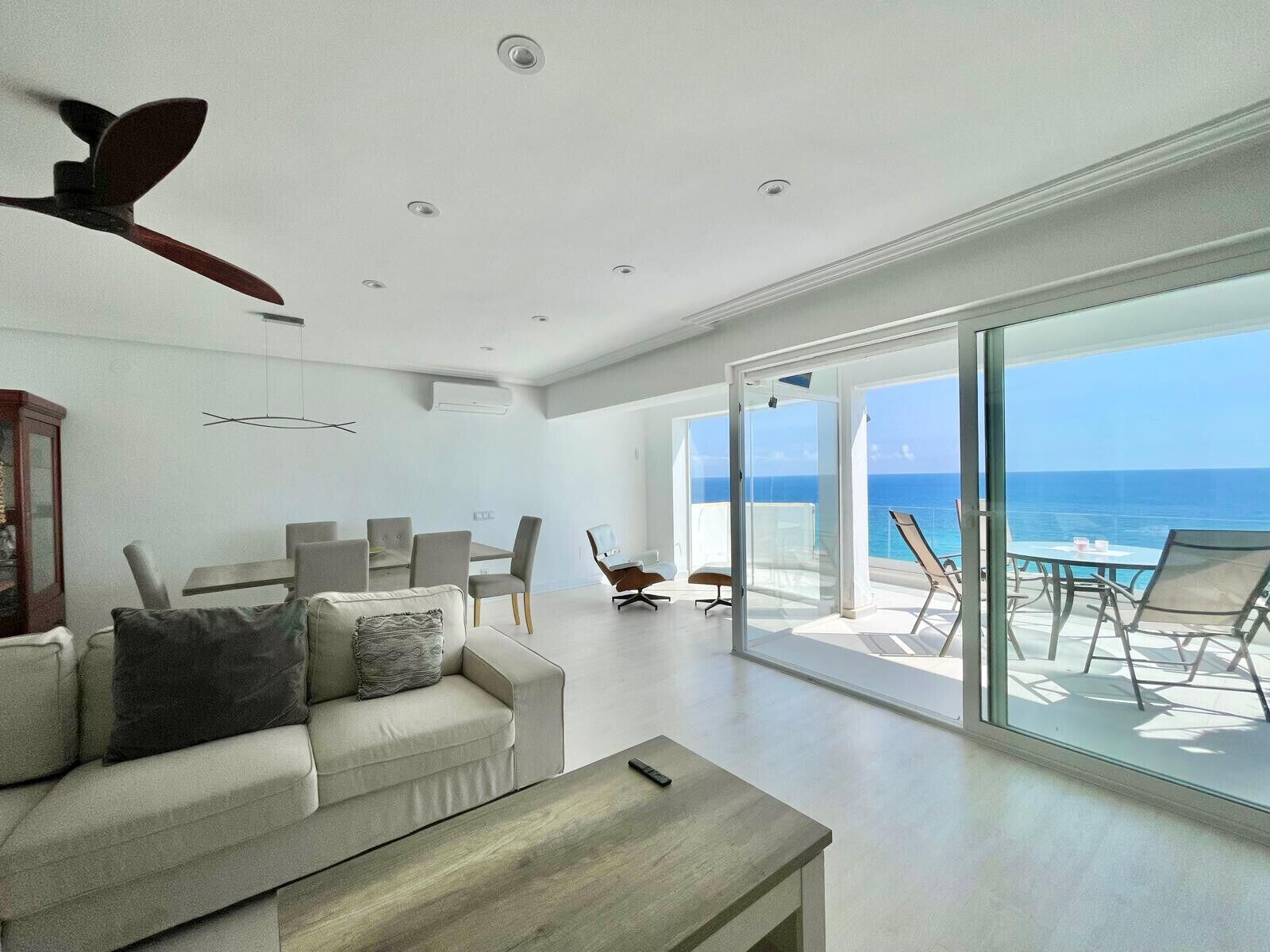 Un salon moderne comprenant un canapé, une table basse et un coin repas. Une grande porte vitrée donne accès à un balcon offrant une vue sur l’océan.