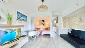 A modern hotel room with a large flat-screen TV, a dining area with a table and chairs, a sofa, and a kitchen area.