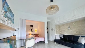 A modern apartment with a glass dining table, a black sofa, a bed with a pink and white patterned bedspread, and a wall with heart-shaped designs.