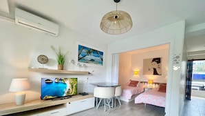 A modern hotel room with a TV, a dining table, two beds, a small kitchen area, and a balcony.