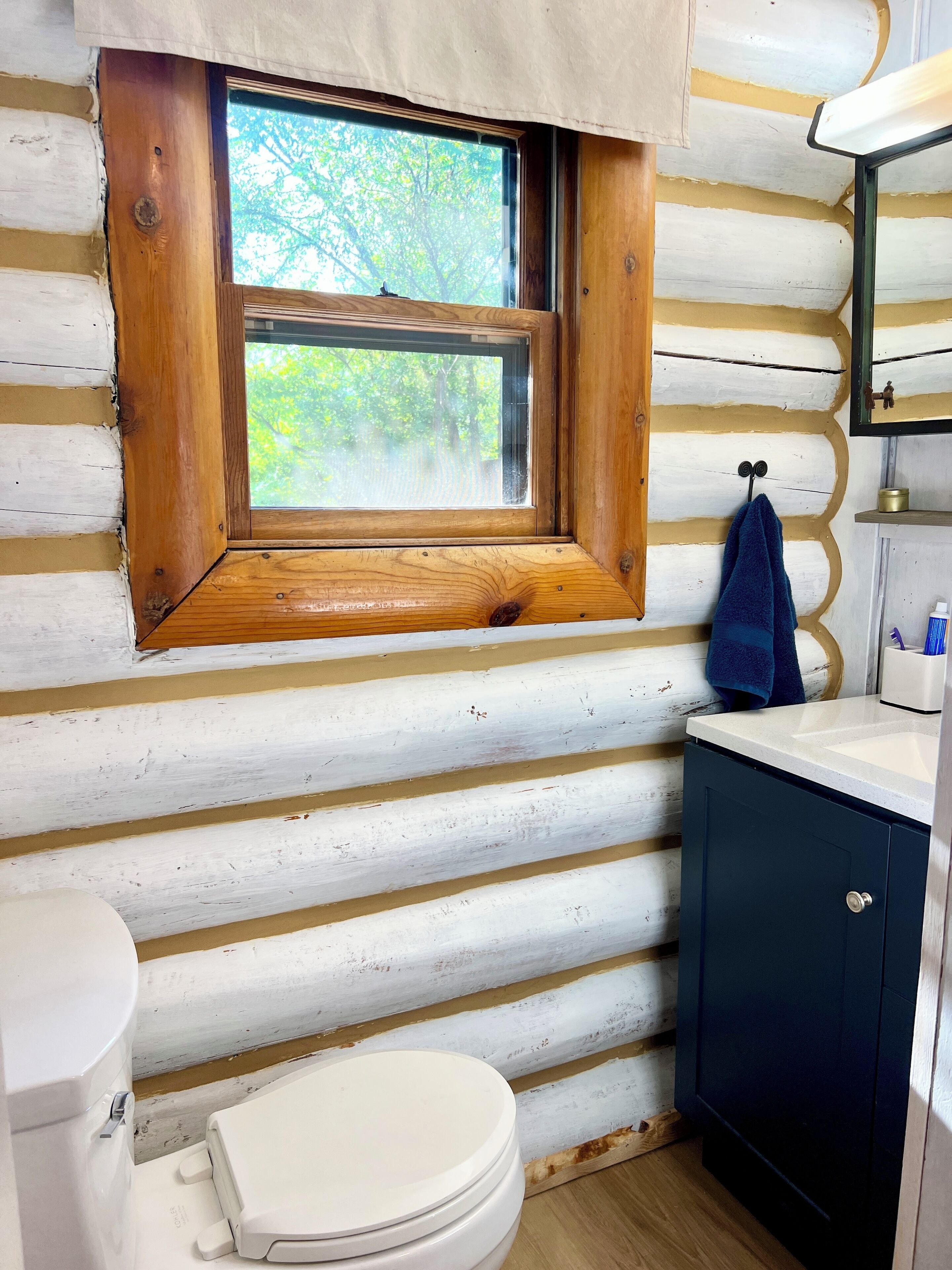 Un baño de estilo cabaña con pared de madera, una ventana con marco de madera, un inodoro blanco, un lavamanos con mueble oscuro y una toalla colgada en un gancho.