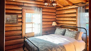 Ein Schlafzimmer in einer Blockhütte mit Bett, einer Kandelaberleuchte, einem Fenster mit Vorhängen und einem Bild an der Wand.