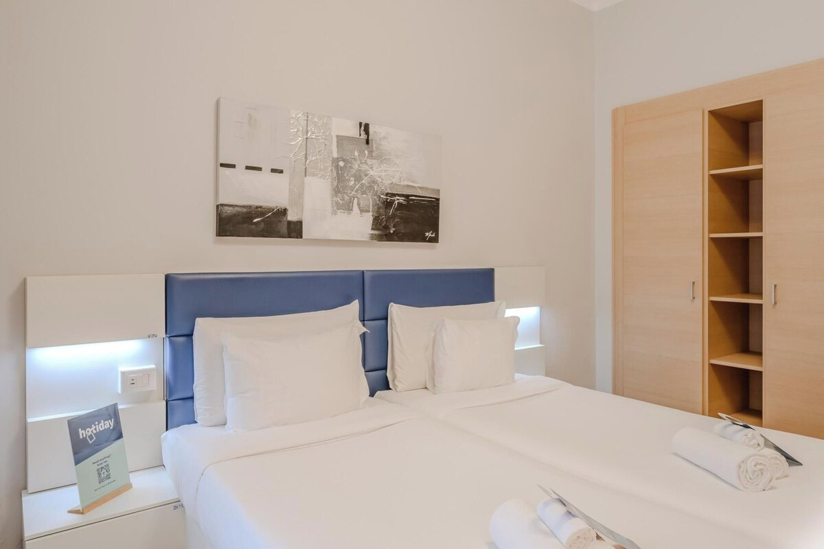 A hotel room with a bed, a nightstand, a wall-mounted artwork, and a wooden wardrobe.