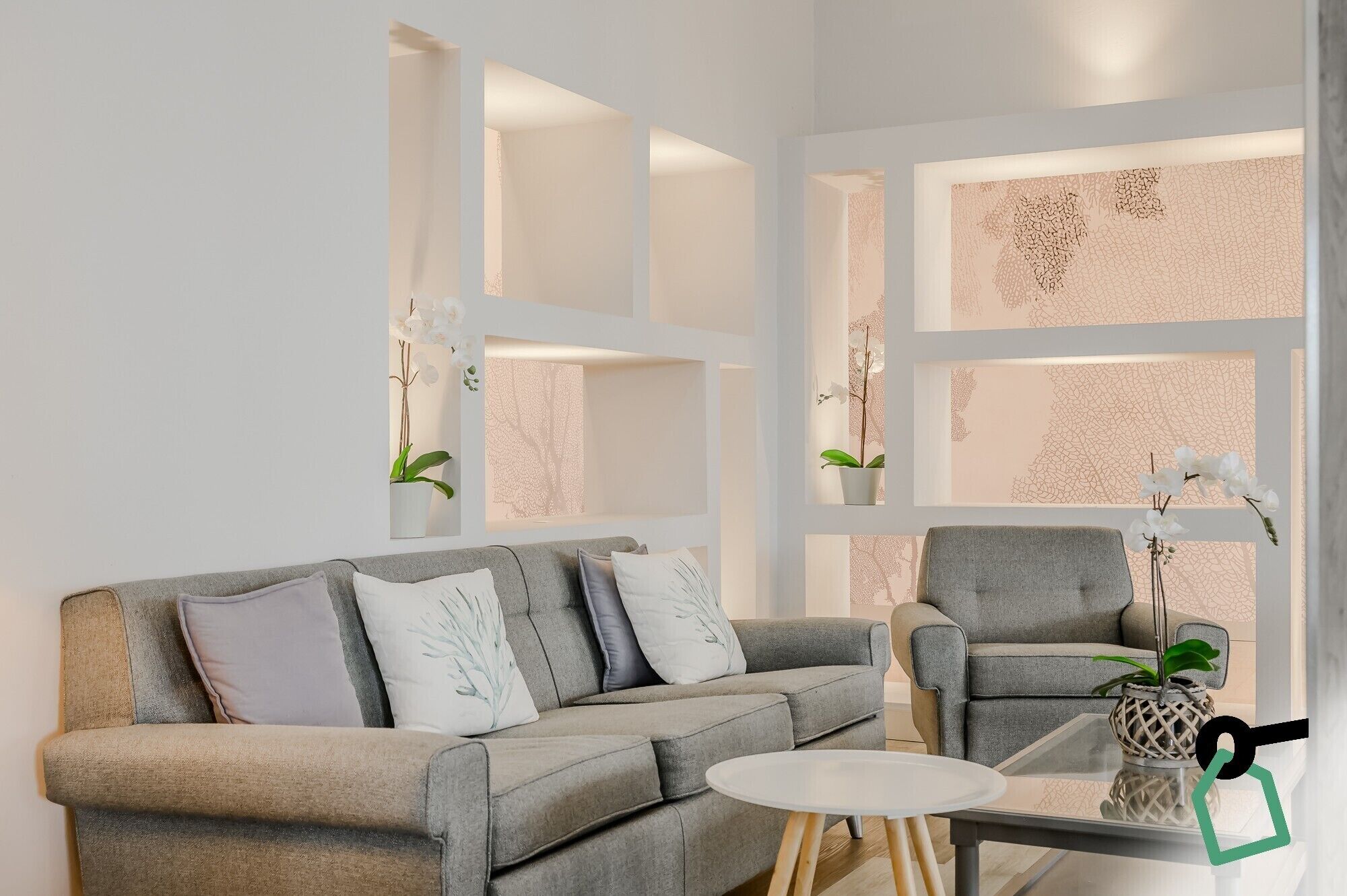 Un salon moderne avec un canapé gris, un fauteuil assorti et une petite table ronde. On y trouve des plantes décoratives et un mur orné d’un motif texturé.