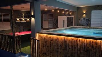 Un área de piscina interior con terraza de madera, mesa de billar y una escalera que lleva a un balcón.