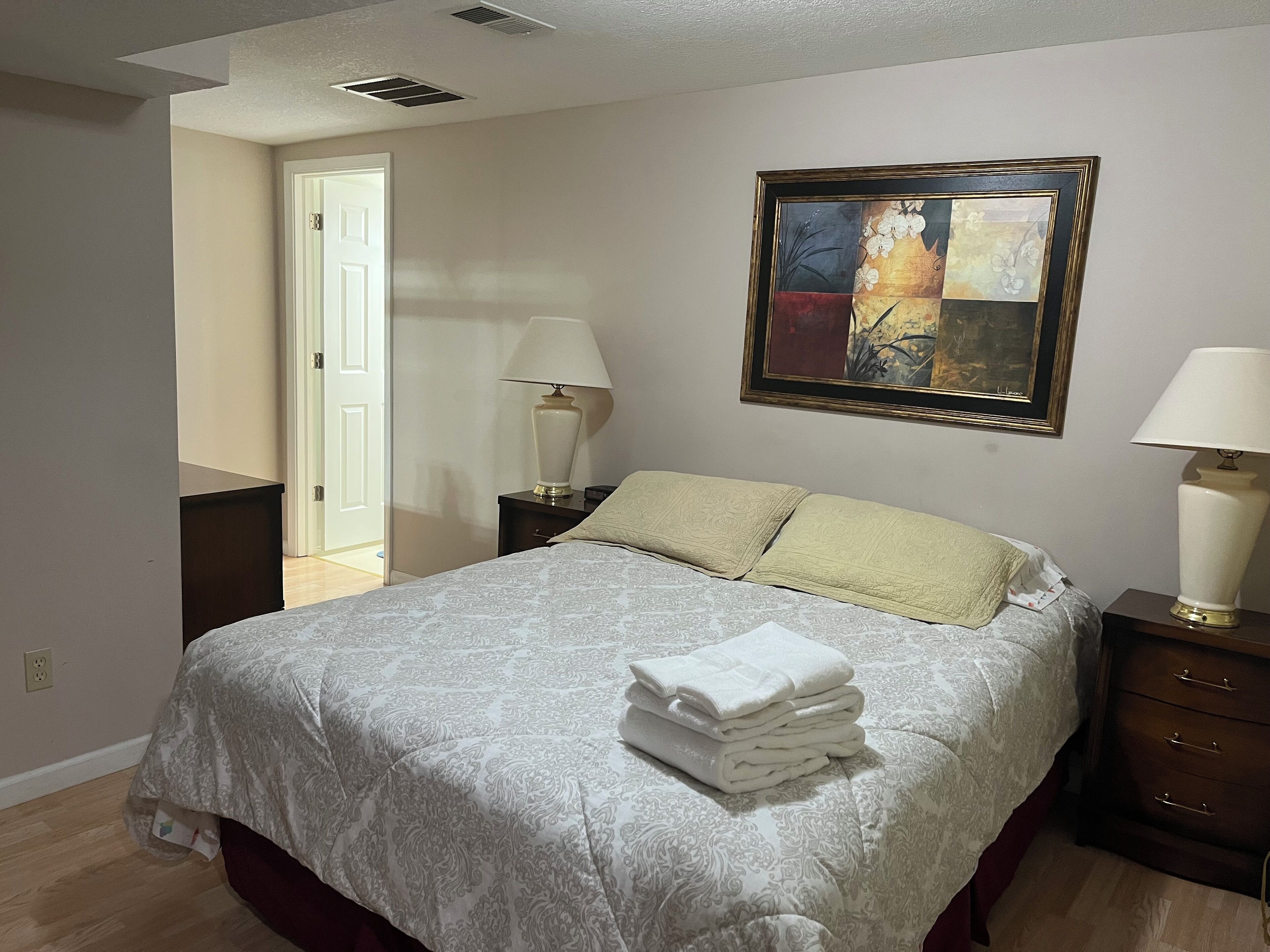A neatly made bed with a patterned bedspread and pillows, a framed painting on the wall, and a lamp on a bedside table.