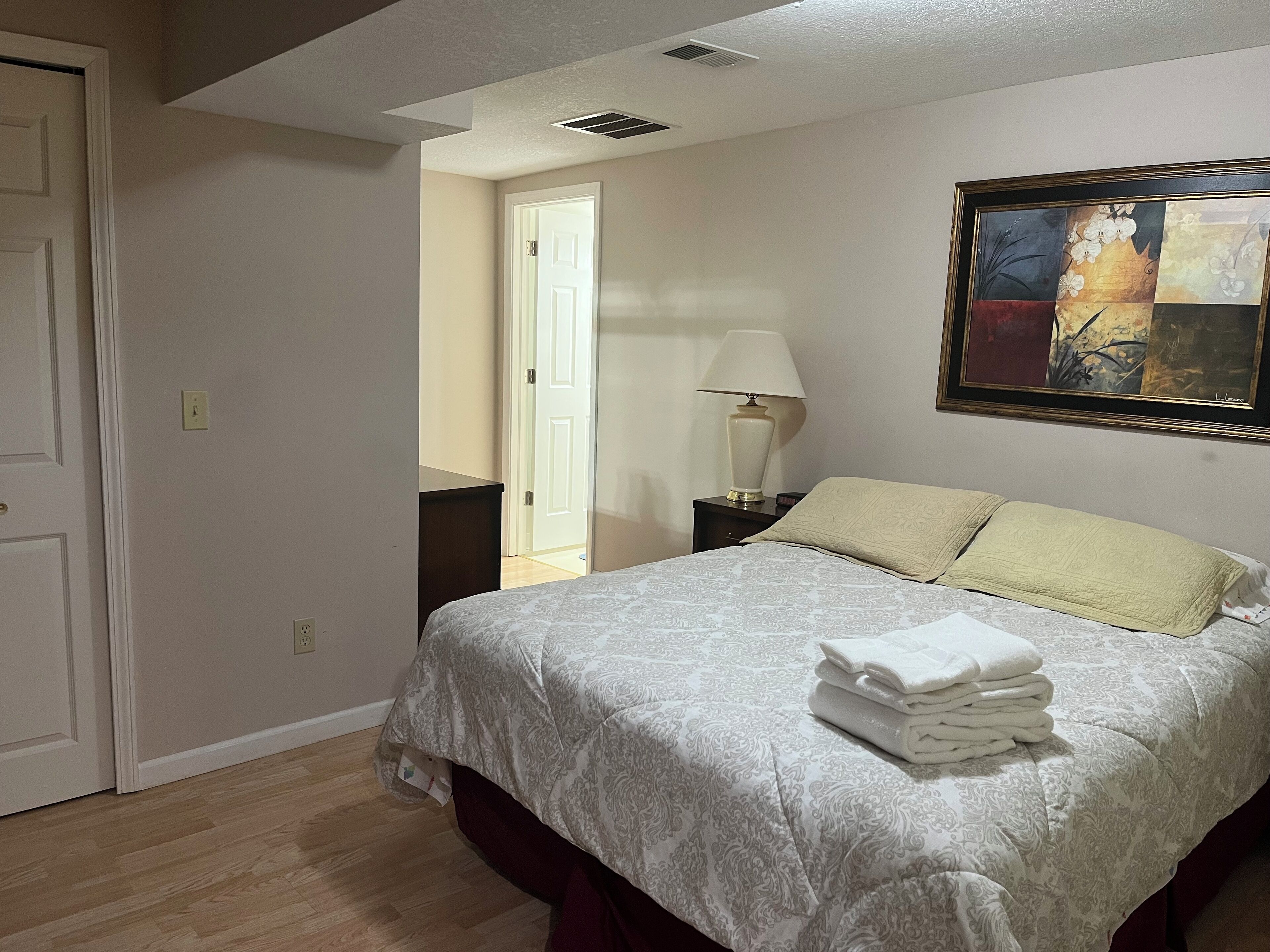 Un dormitorio con cama, mesita de noche, lámpara y un cuadro en la pared.