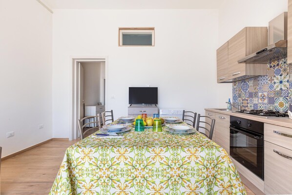 Un coin repas avec une table dressée pour quatre personnes, un téléviseur placé au-dessus de la table, et un espace cuisine doté d’un dosseret carrelé et de meubles en bois.