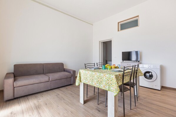 A small, well-lit room with a sofa, a dining table with chairs, a washing machine, and a television.