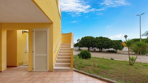 Ein gelbes Gebäude mit einer Treppe, die zu einer Tür führt, einer gefliesten Terrasse und Blick auf eine Straße mit Bäumen und einer Palme.