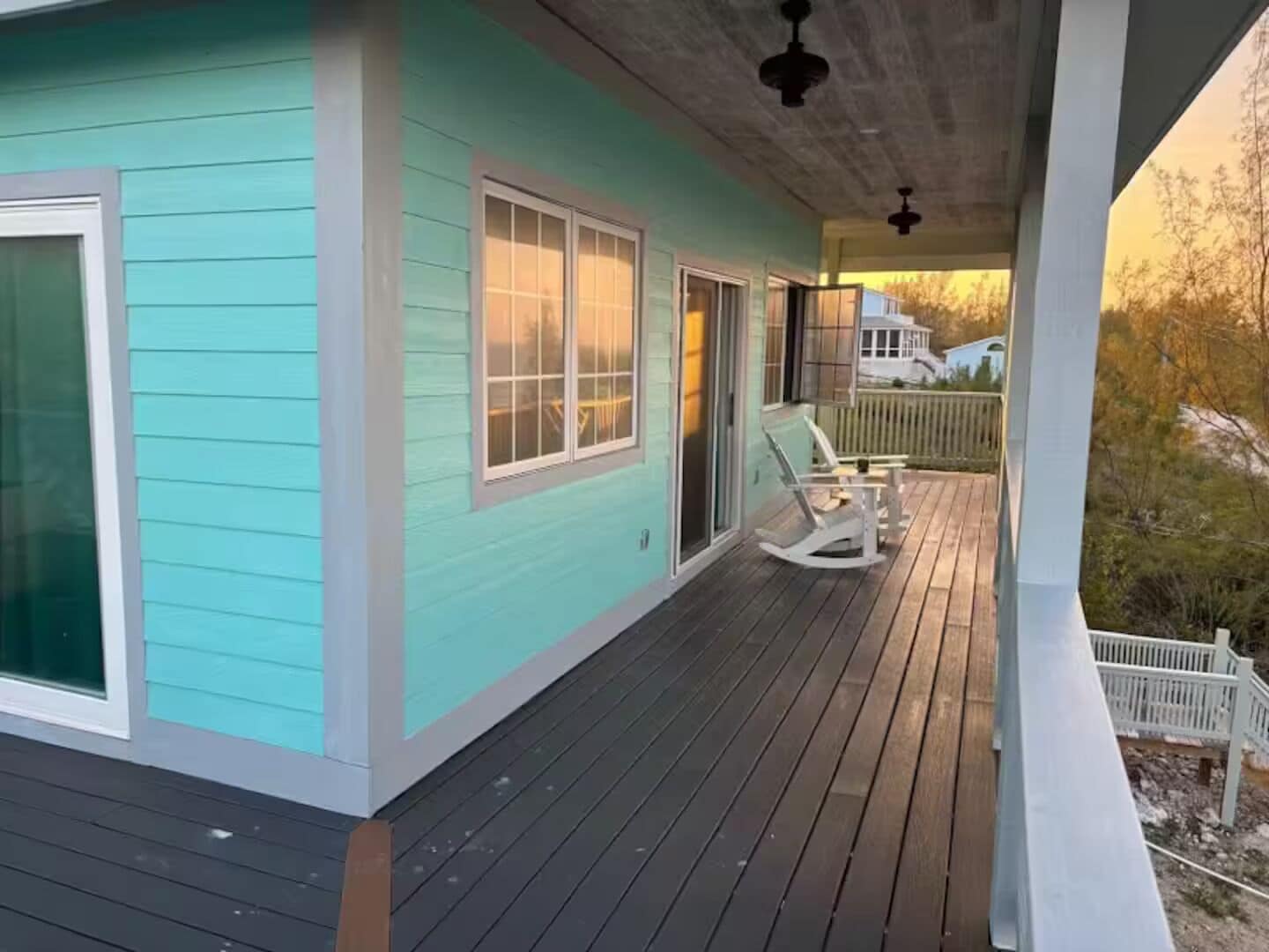 Uma casa turquesa com varanda de madeira, uma cadeira de balanço e cerca branca.