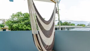 A hammock hanging on a balcony with a view of buildings and trees in the background. - Home for 15 Guests with Private Pool and Pool Table (Aguadilla)