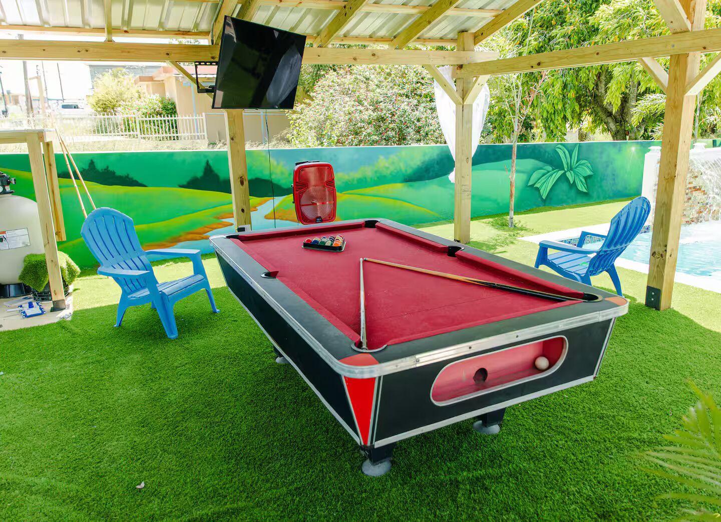 A pool table with a red felt surface, surrounded by two blue chairs, under a wooden pergola with a TV mounted above.
