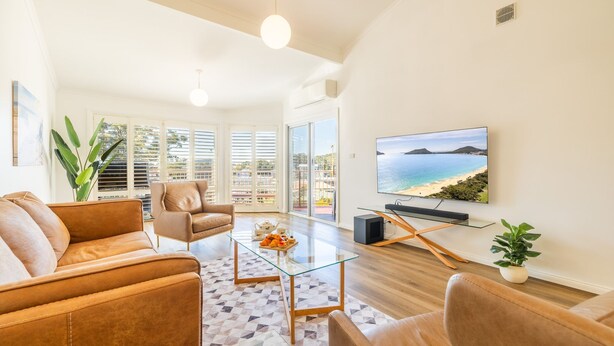 A modern living room with leather sofas, a glass coffee table, and a large flat-screen TV displaying a beach scene. - Tomaree Mews 5, 18 Tomaree St - Stunning townhouse in the heart of town (Nelson Bay)