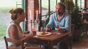 A couple dining at a rustic outdoor table with string lights, surrounded by large windows and greenery.
