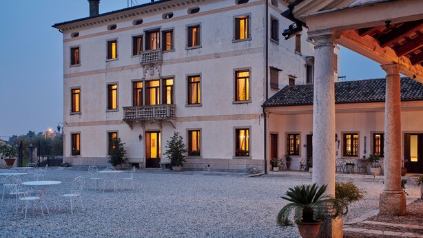 A building with multiple windows and a balcony, surrounded by a gravel courtyard with outdoor seating.