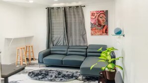 Un salon moderne avec un canapé d’angle gris foncé, un tapis rond, des tabourets en bois, une plante en pot et une grande œuvre d’art encadrée accrochée au mur.
