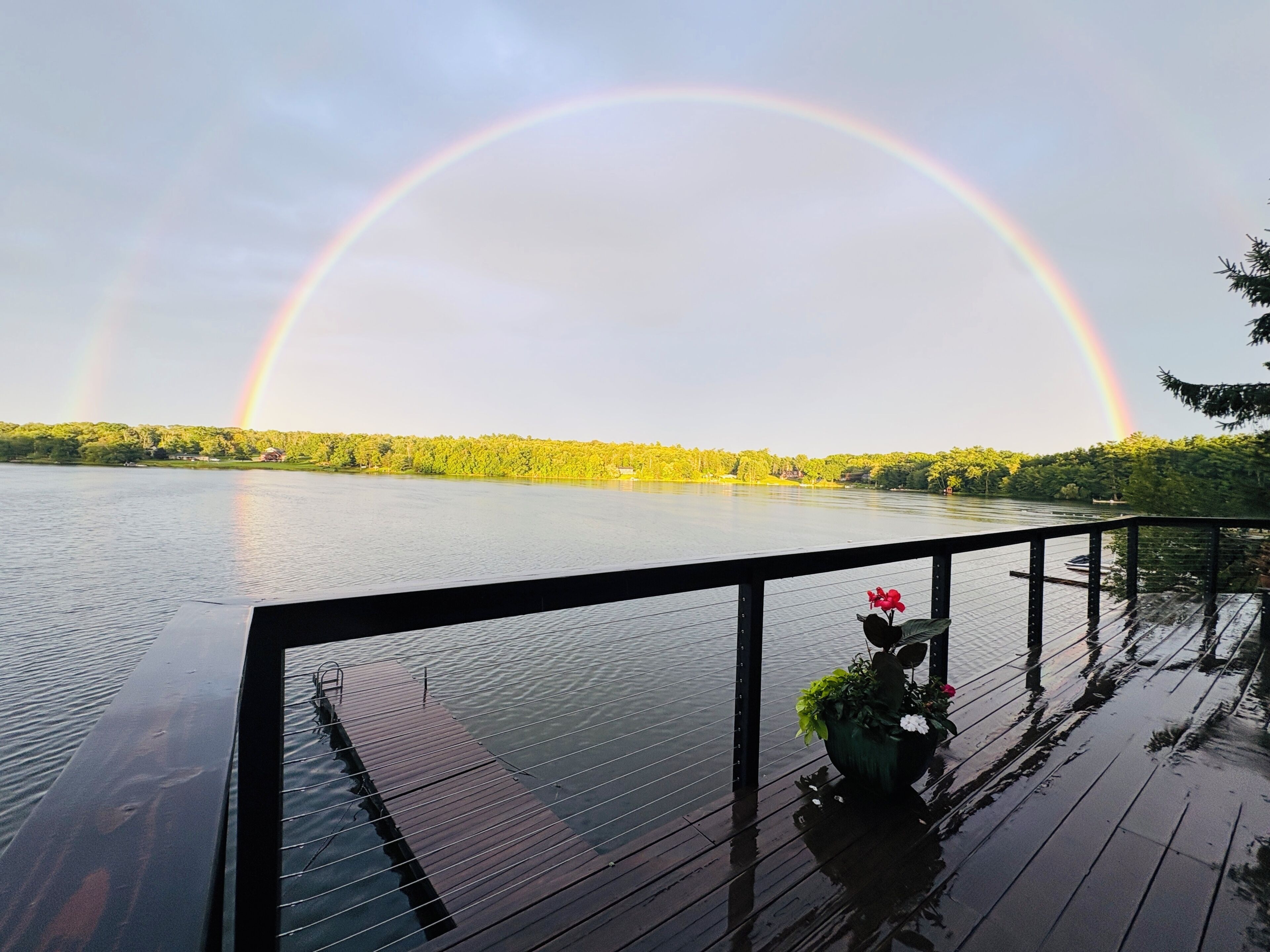 A wooden deck with a railing, overlooking a calm lake with a rainbow in the background.