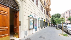 A street view with a building featuring a large wooden door and a metal awning, storefronts with signage, and parked cars. - Cola di Rienzo apartment (Rome)