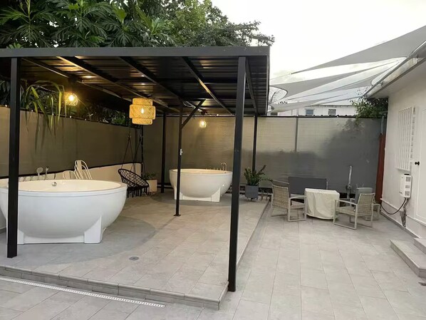 A modern outdoor bathroom with two large tubs, a covered seating area, and potted plants.