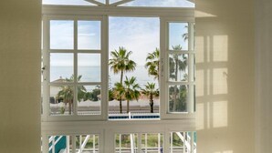 A room with a large bay window overlooking a pool and palm trees, with a view of the sea.