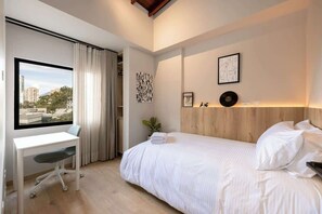 A modern hotel room with a large window, a desk, a bed, and a wooden headboard.