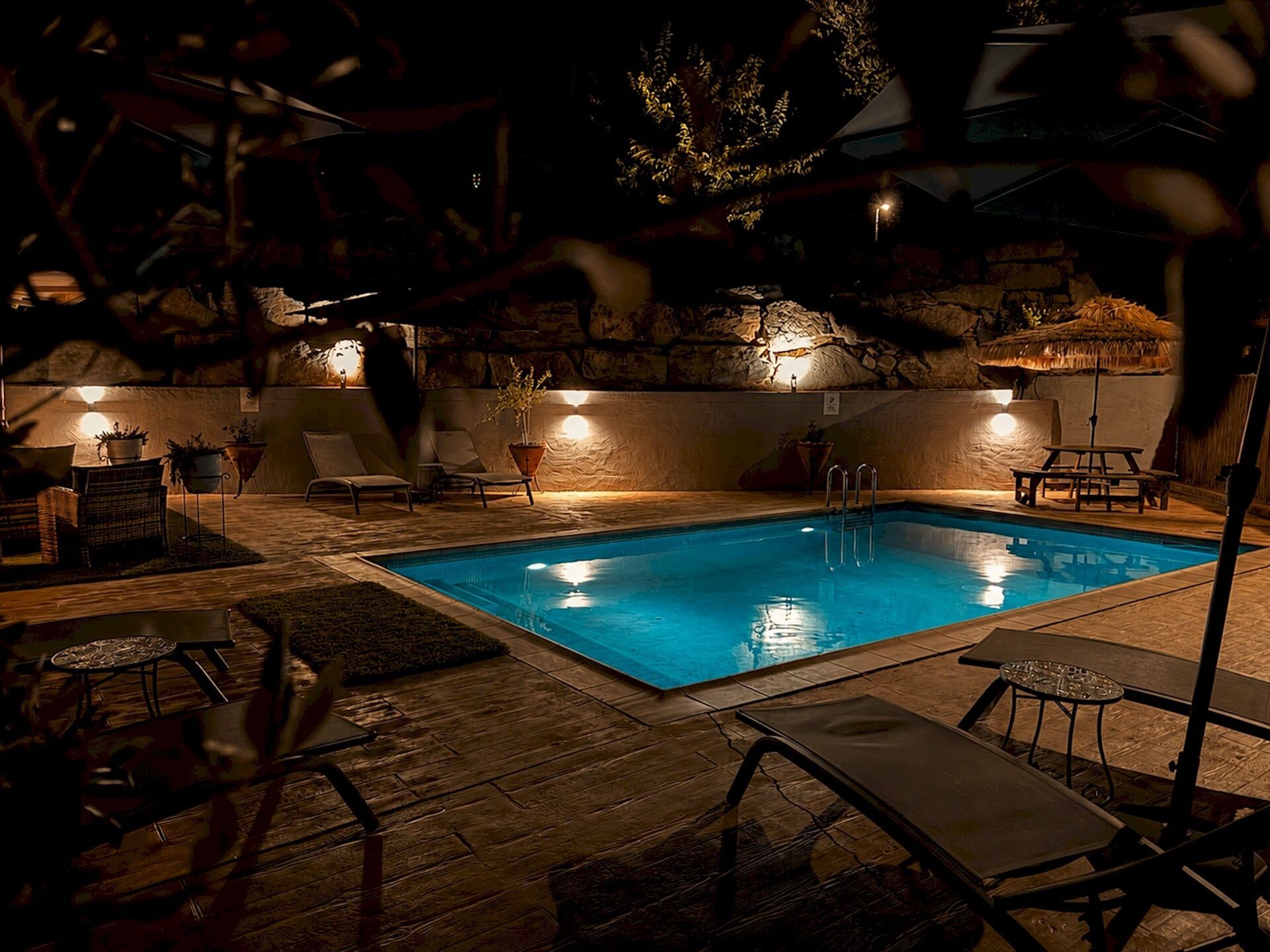 Ein Swimmingpool mit Liegestühlen und Blumentöpfen, umgeben von Steinmauern und warmer Beleuchtung.