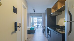 Un pasillo estrecho que conduce a un dormitorio con una cama, una mesita y una silla. A la derecha, hay una kitchenette con estufa y refrigerador.
