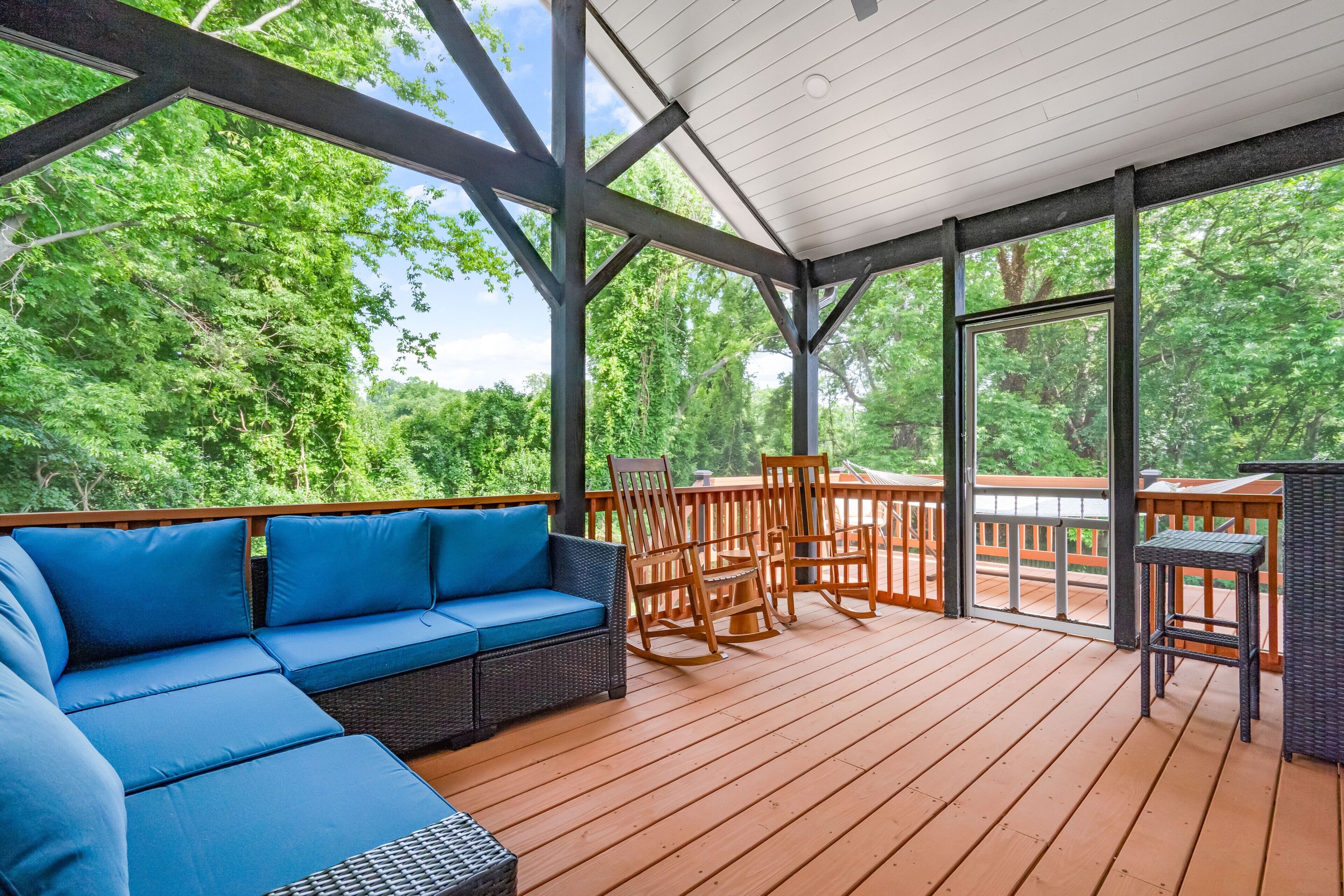 Een overdekt terras buiten met een blauwe bank, houten stoelen en een tafel.