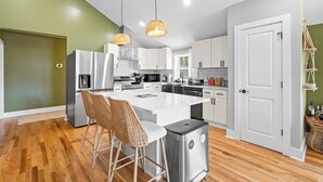 A modern kitchen with white cabinets, a central island with bar stools, stainless steel appliances, and a hanging light fixture. - Boho 5BR Modern Villa•Hot Tub•Grill•Patio•Backyard (Raleigh)