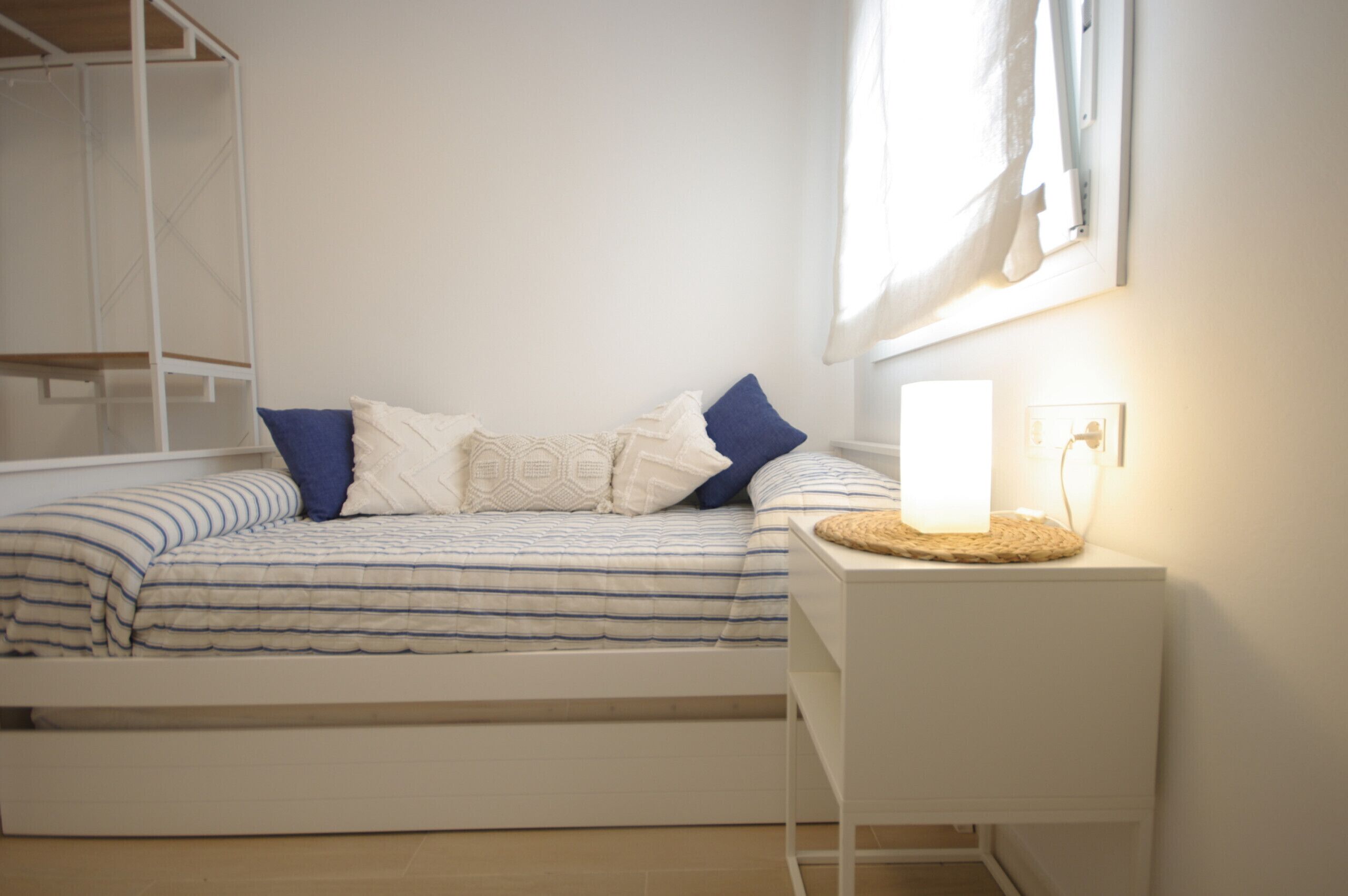 Un letto con copriletto a righe, due cuscini, un comodino con una lampada e una finestra con tende.