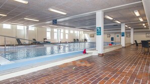 Una piscina interior con suelo de baldosas, techo transparente con luces y algunas personas nadando.