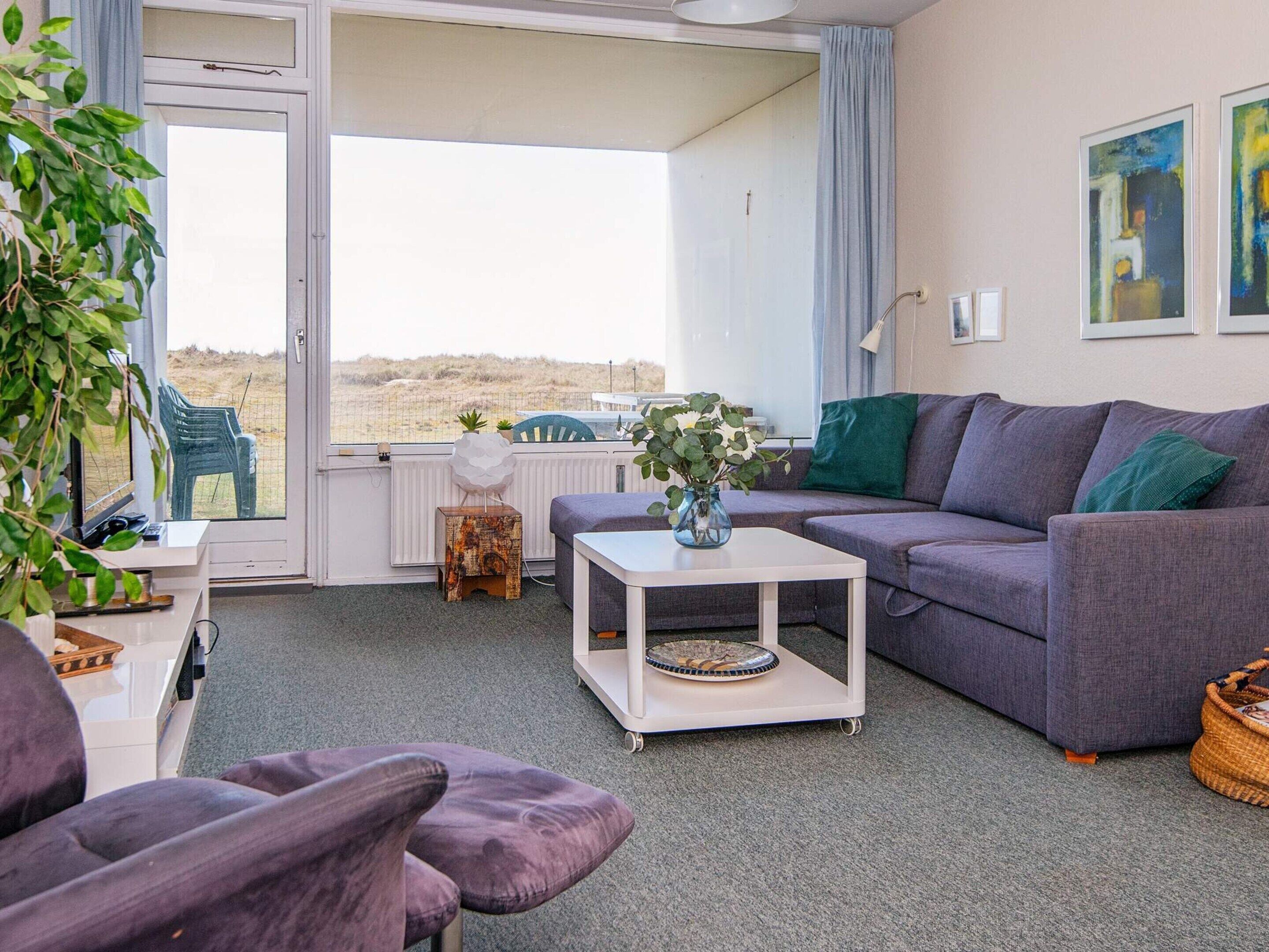 Ein gemütliches Wohnzimmer mit einem grauen Sofa, einem weißen Couchtisch und einem violetten Sessel. Ein Fenster bietet Blick auf den Strand.