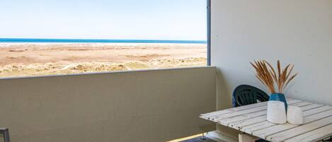 Un balcón con vistas a la playa, una mesa con un florero y una silla, y una pared con un cartel pequeño.