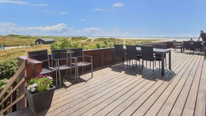 Terraza de madera con asientos exteriores, con vista a una playa y dunas.