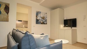 A modern living room with a grey sofa, a flat-screen TV mounted above a white cabinet, and a kitchen area in the background.