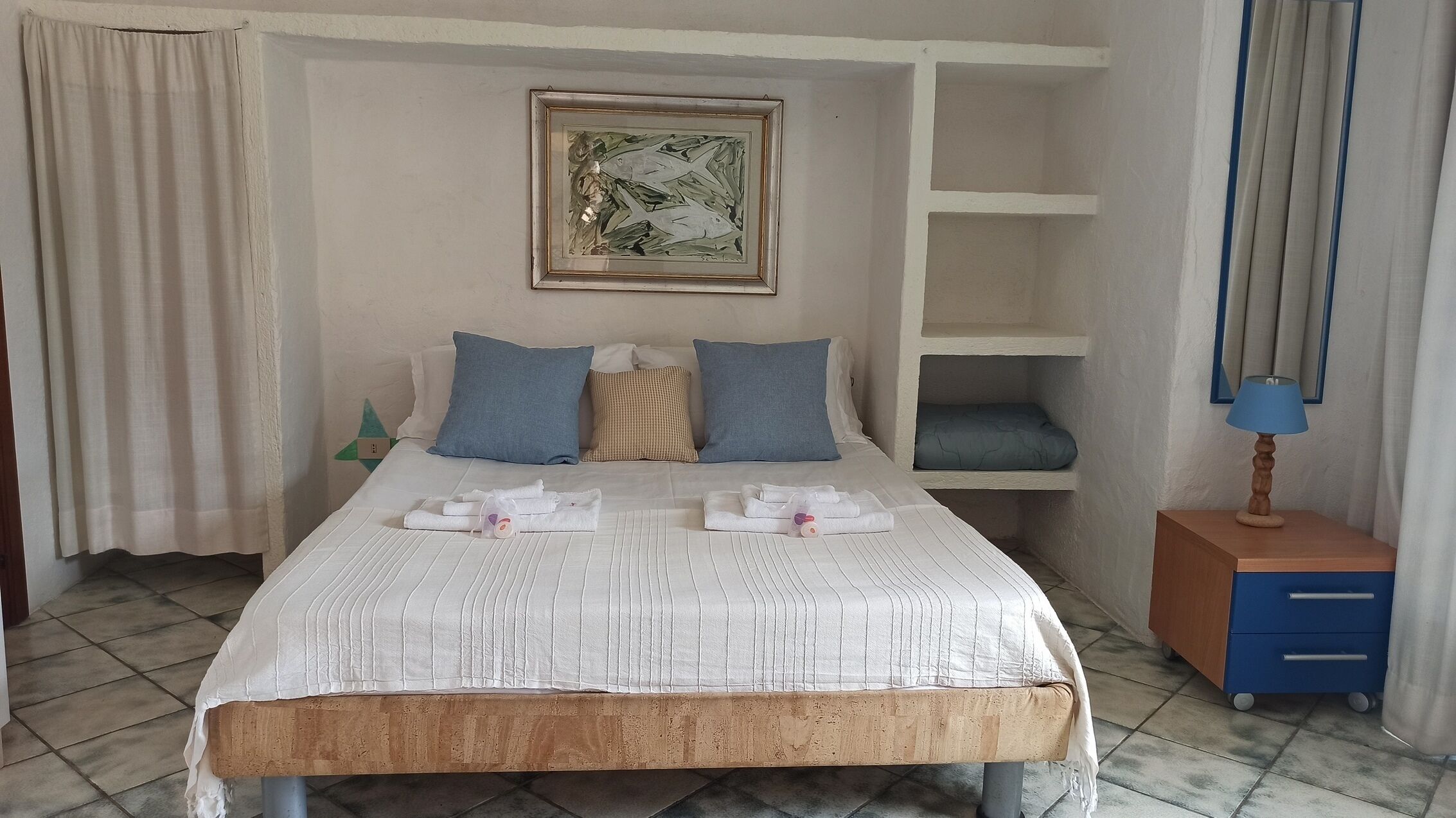A neatly made bed with pillows, a framed artwork above it, and a small blue table with a lamp beside the bed.