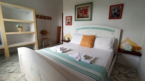 A neatly made bed with white and green striped bedding, a wooden nightstand with a clock and a vase of flowers, and a shelf with a vase of flowers. - Domos De Badas - Trilocale (Porto Ottiolu)