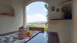 A table with a jar labeled "SUGAR Z" and a lemon, a cup, and a spoon on a tablecloth. An archway leads to a view of a landscape with mountains and a body of water. - Domos De Badas - Trilocale (Porto Ottiolu)