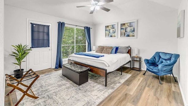 Un dormitorio con cama, ventilador de techo, ventana con cortinas, una silla azul y arte en la pared.